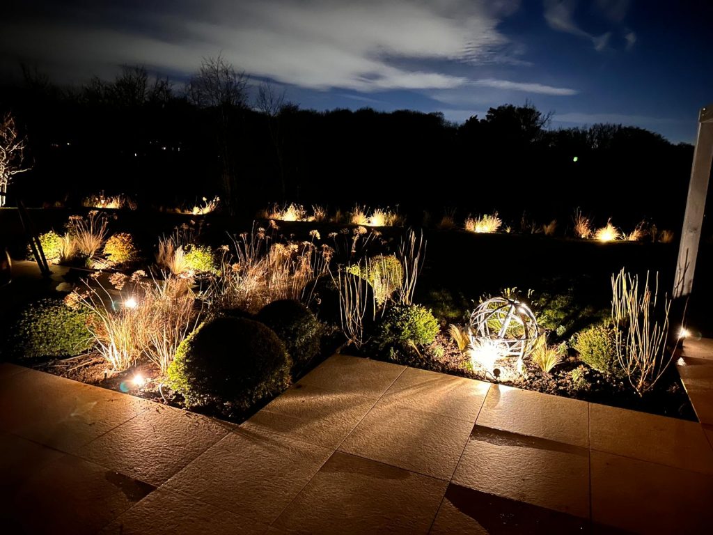 Planted garden border lit at dusk with ornamental grasses and a sphere feature in Kent